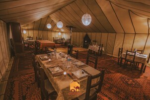 Una carpa con un área de comedor dispuesta en su interior. Mesas dispuestas con sillas, y lámparas colgantes arriba.