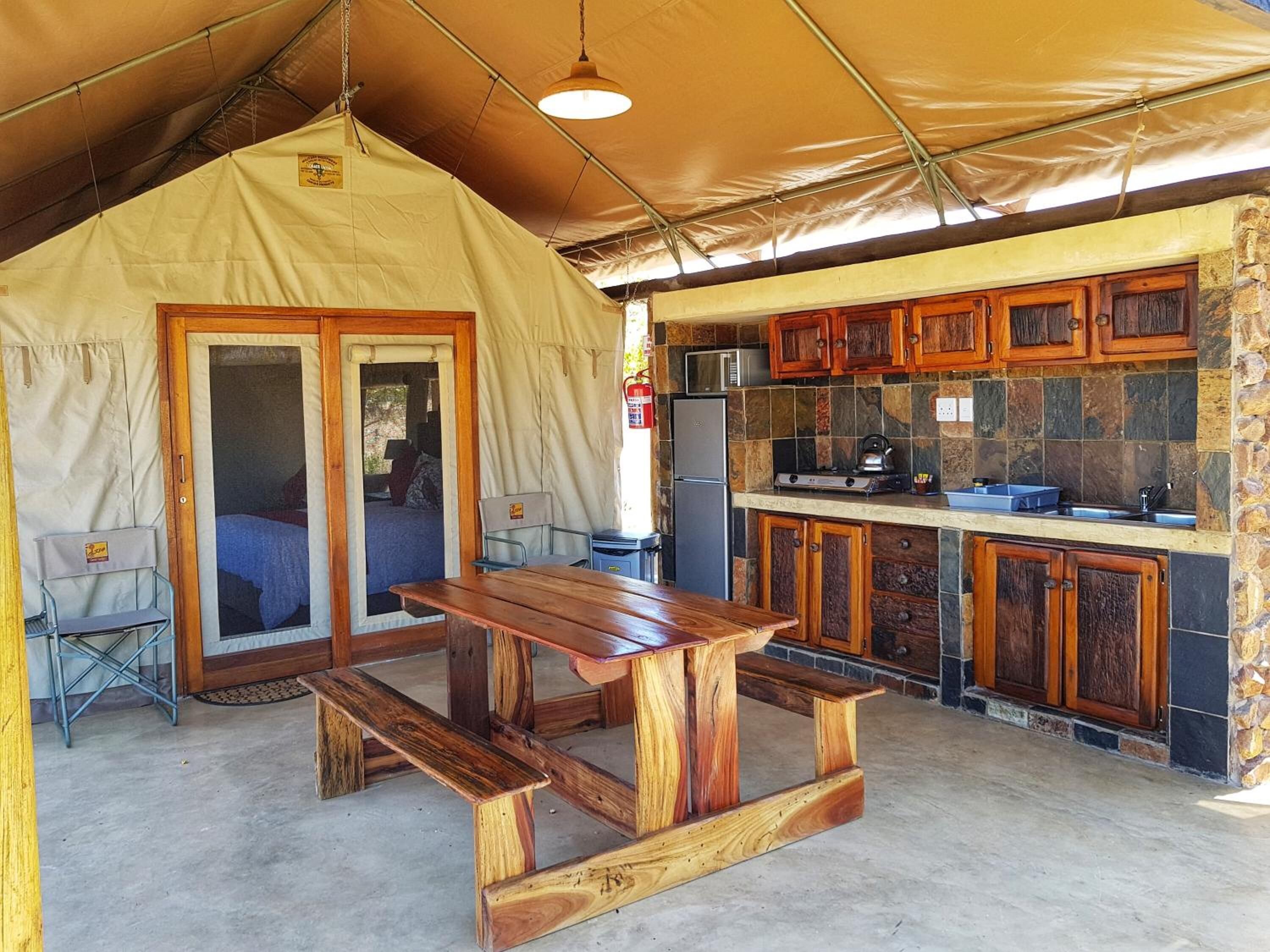 A tented accommodation with a wooden table and benches, a kitchenette with stone accents, and a view of the outdoors.