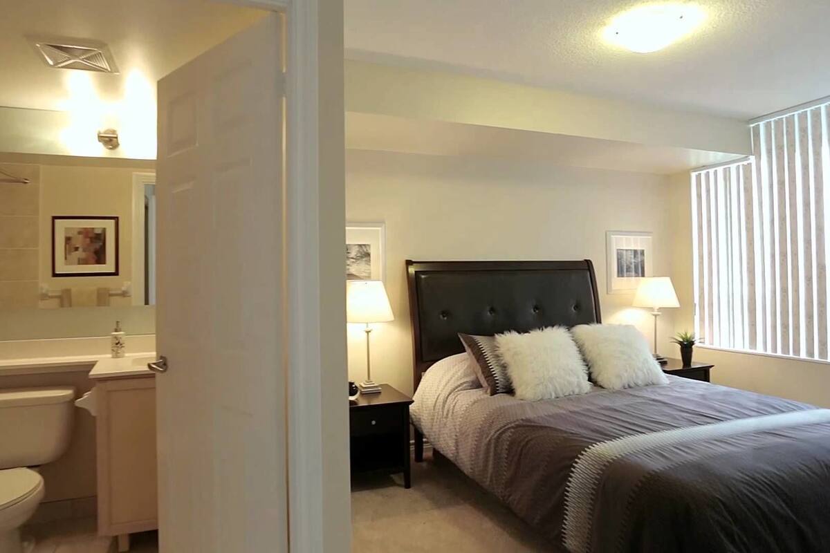 Une chambre à coucher comprenant un lit, des tables de chevet, une lampe, une fenêtre avec des stores, ainsi qu’une salle de bain avec un lavabo et des toilettes.