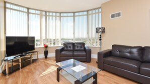 A modern living room with a large flat-screen TV, two leather sofas, a glass coffee table, and large windows with blinds. - NAPA Furnished Suites Square One (Mississauga)