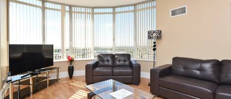 A modern living room with a large flat-screen TV, two leather sofas, a glass coffee table, and large windows with blinds.