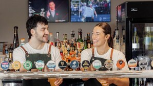 Dos empleados de bar detrás de una barra con varias grifas de cerveza y un refrigerador.