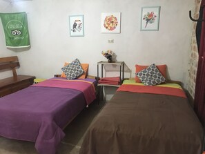 A room with two beds, each with a different colored bedspread and patterned pillows. There is a small table with a vase of flowers, and a green bag hanging on the wall.