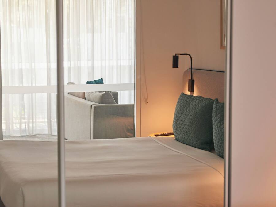 Ett hotellrum med en säng, en stol, ett skrivbord och ett fönster med tunna, genomskinliga gardiner.