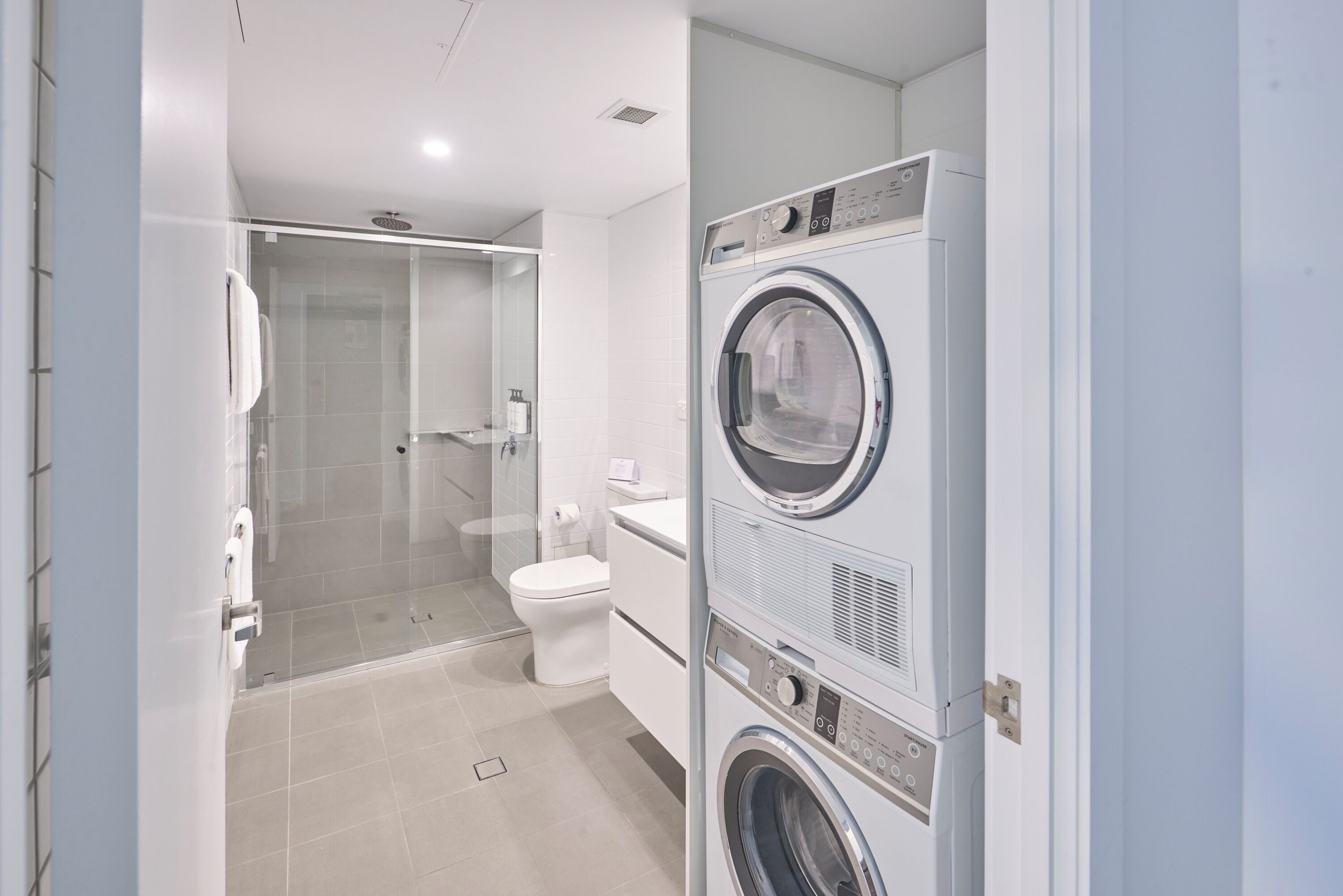 A modern bathroom with a glass shower enclosure, a toilet, and stacked laundry machines.