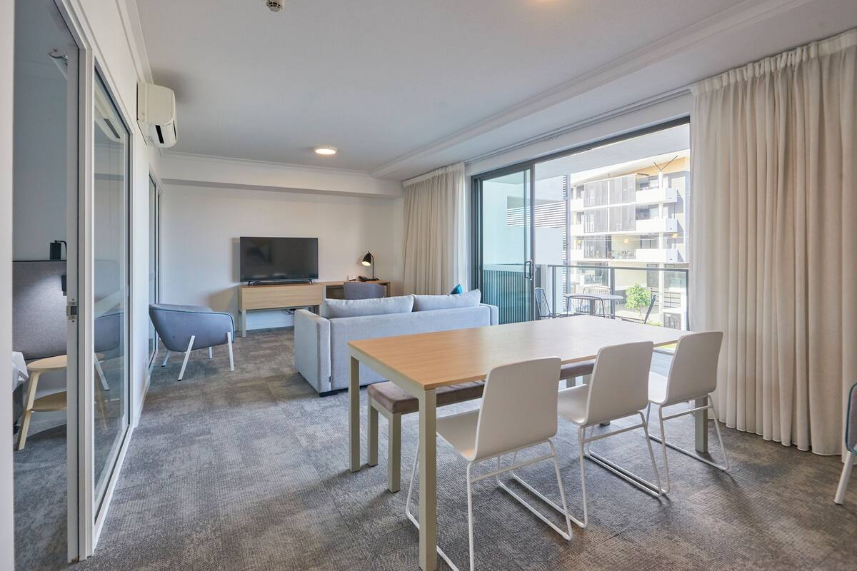 Quarto de hotel moderno com área de refeições, sofá, TV e varanda com cortinas.