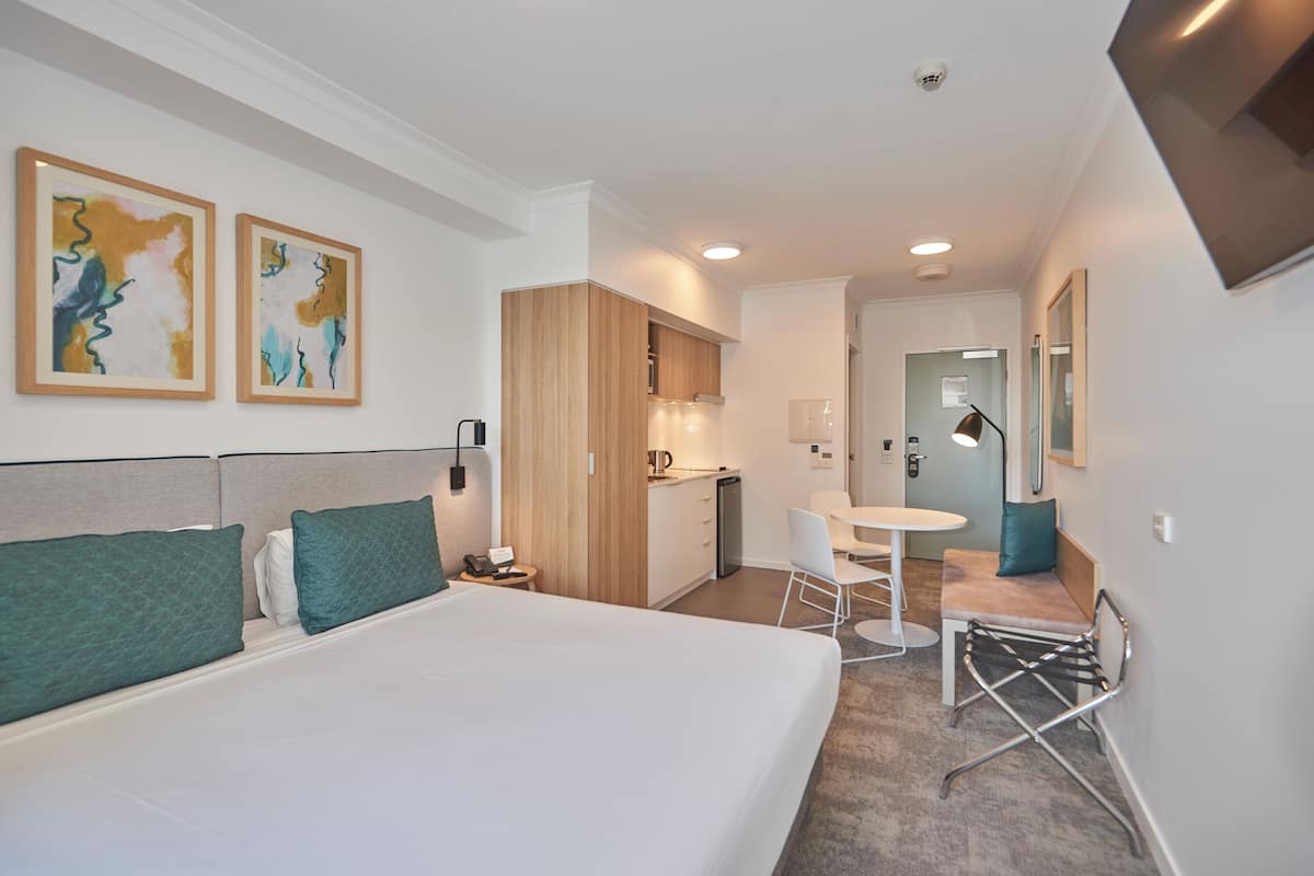 Quarto de hotel com cama grande, escrivaninha, cadeira, pequena cozinha e duas pinturas abstratas na parede.