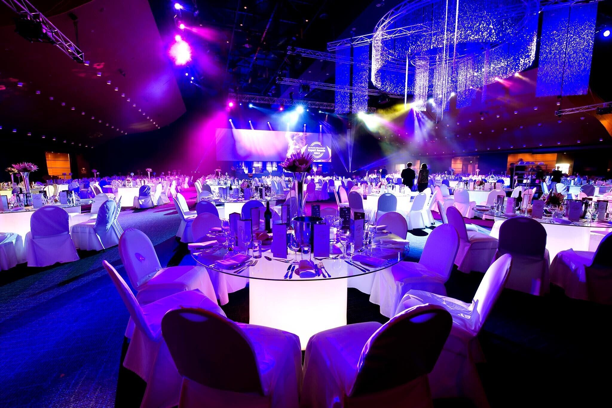 A banquet hall with round tables, white chairs, and a stage with lighting.