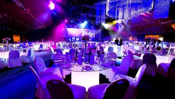 A banquet hall with round tables, white chairs, and a stage with lighting.