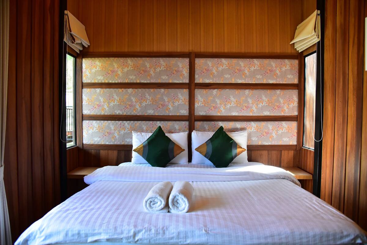Habitación de hotel con cabecera de madera, cortinas con estampado floral, dos almohadas, una cama con toallas enrolladas y un banco de madera.