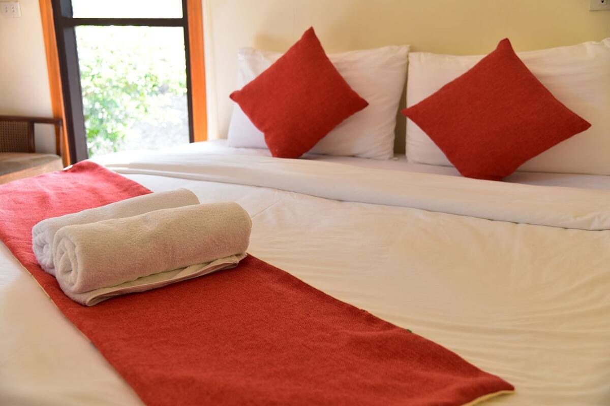 Una cama bien tendida con sábanas blancas y almohadas rojas, una toalla roja sobre la cama, y una ventana con vistas a zonas verdes.