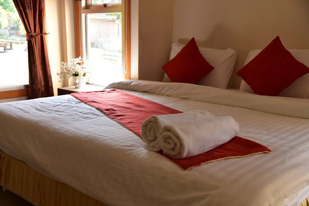 Una cama bien hecha con ropa de cama roja y blanca, una toalla enrollada y un florero con flores blancas sobre una mesita de noche.