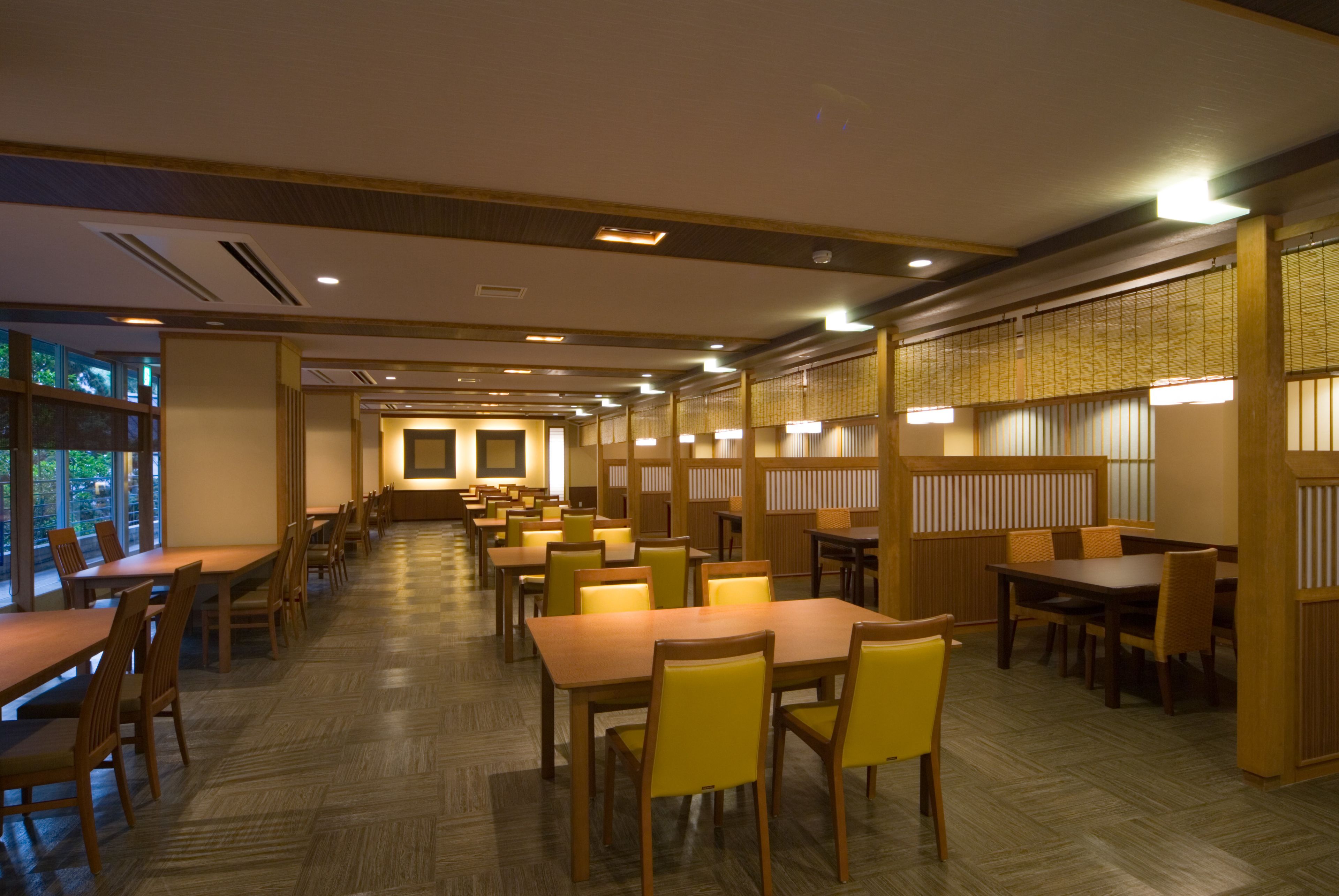 A spacious dining area with multiple tables and chairs, wooden flooring, and a view of greenery through large windows.