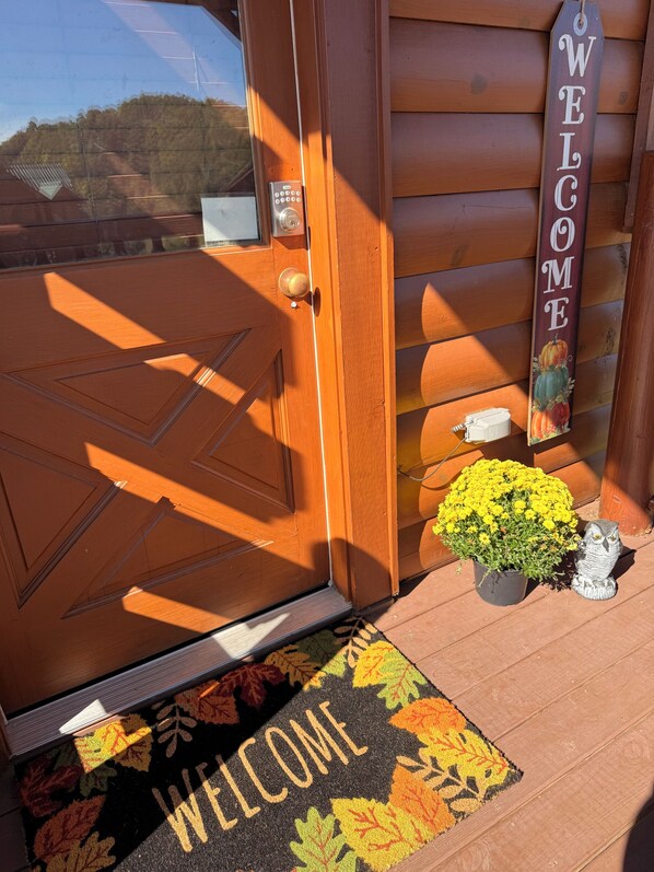 A wooden door with a keypad lock, a welcome mat with autumn leaves, a potted plant with yellow flowers, and a wooden sign that says "WELCOME".