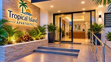 The entrance to Tropical Island Apartment Hotel with a reception desk, indoor plants, and a staircase leading to the interior.