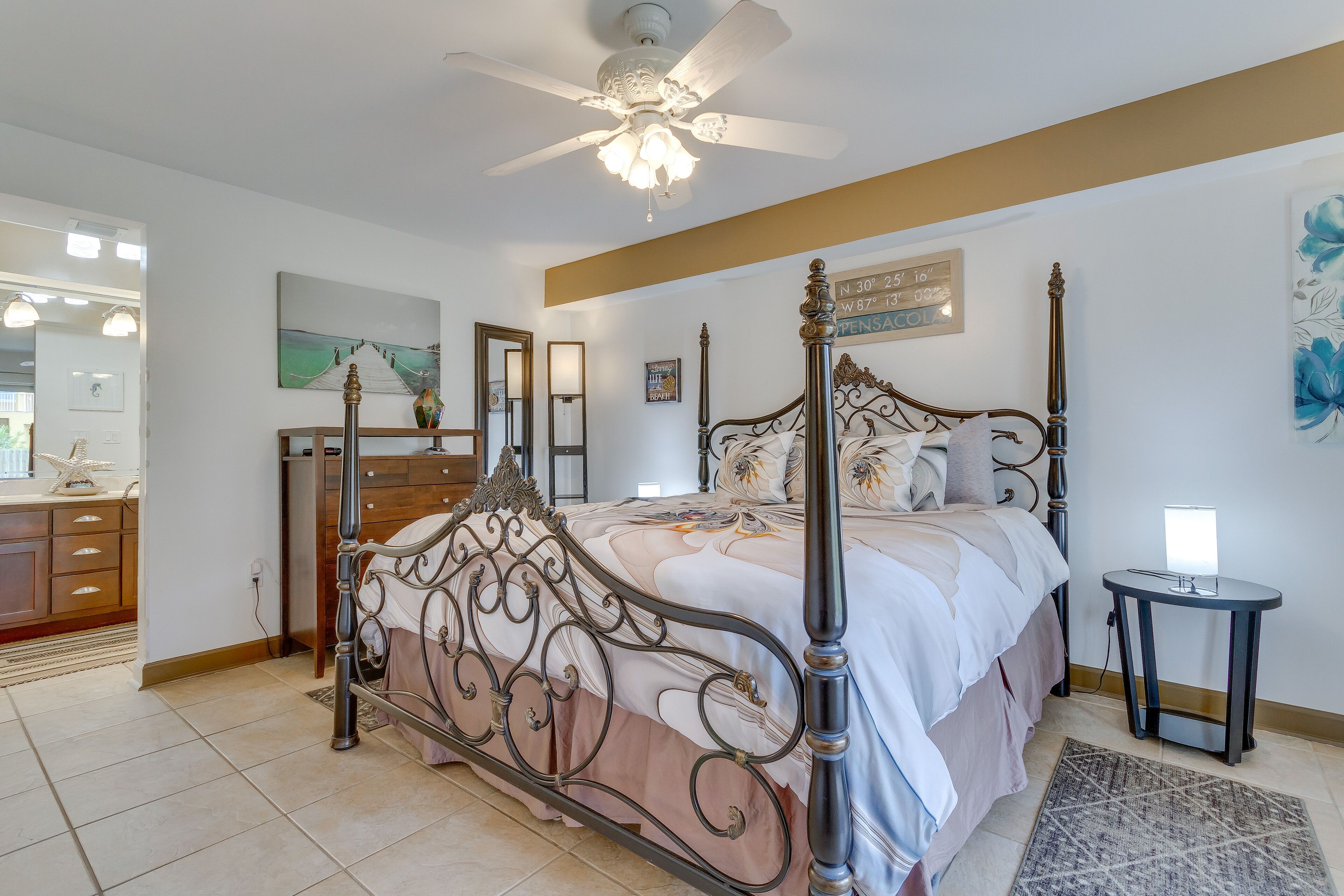 A bedroom with a four-poster bed, a bedside table, a small round table, a ceiling fan, and a painting on the wall.