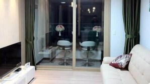Uma sala de estar moderna com um sofá, uma mesa de centro e uma televisão. Há uma porta de vidro deslizante que dá para uma varanda com uma cadeira e uma mesa.