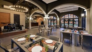 Un restaurante espacioso con techos altos, ventanas arqueadas y un escenario con instrumentos musicales. Las mesas están dispuestas con platos blancos y posavasos rojos.