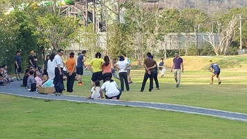 A group of people gathered in an outdoor setting with a grassy area, trees, and a structure in the background.