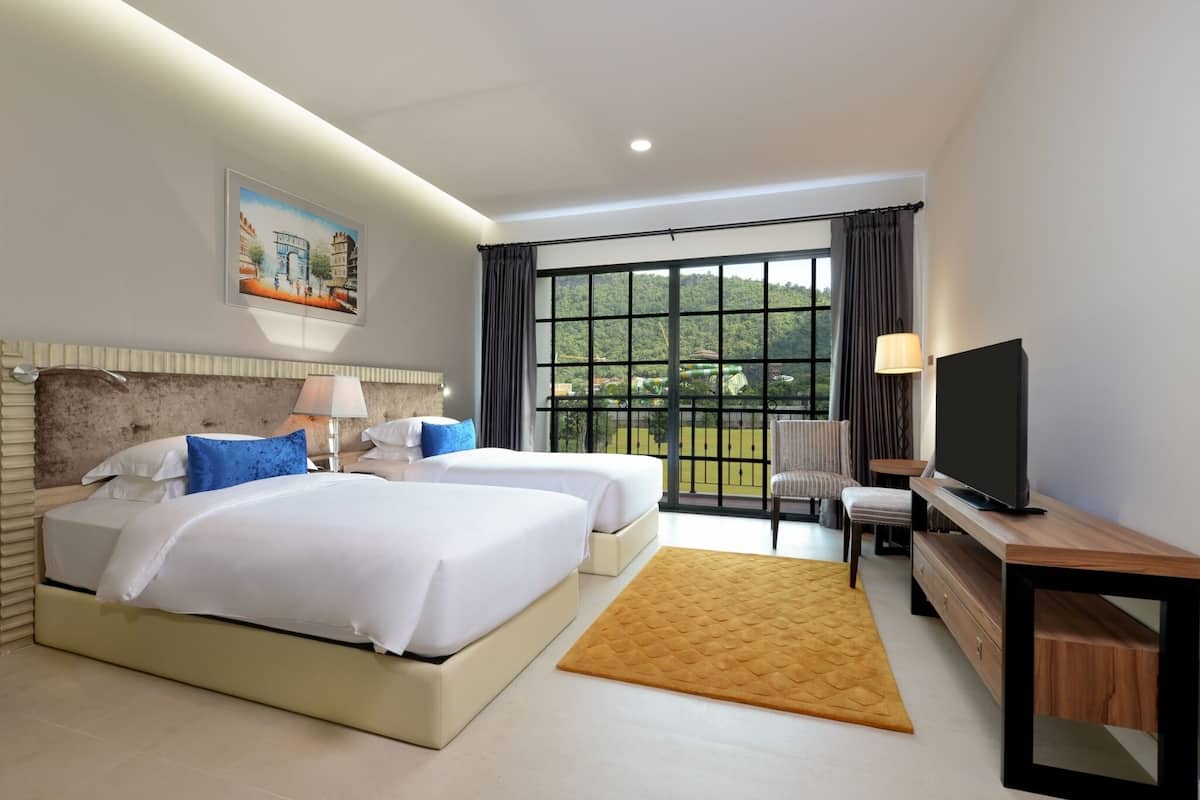 A hotel room with two beds, a TV, a desk, and a large window with a view of greenery.