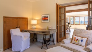 Une pièce avec un bureau en bois, une chaise blanche, un fauteuil rembourré et une vue sur l’extérieur par une porte ouverte.