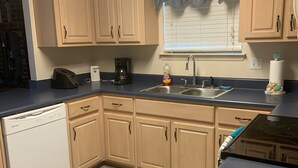 A kitchen with light wooden cabinets, a black countertop, a dishwasher, a coffee maker, and a window with a blue valance. - Cozy, comfortable and convenient (Pensacola)