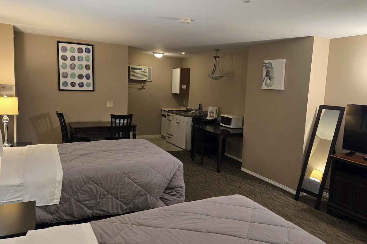 A hotel room with two beds, a desk, kitchenette, TV, and artwork on the walls.