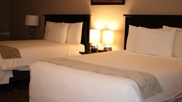 A hotel room with two beds, a nightstand with a lamp, and a painting on the wall. - Red Coat Inn (Fort Macleod)