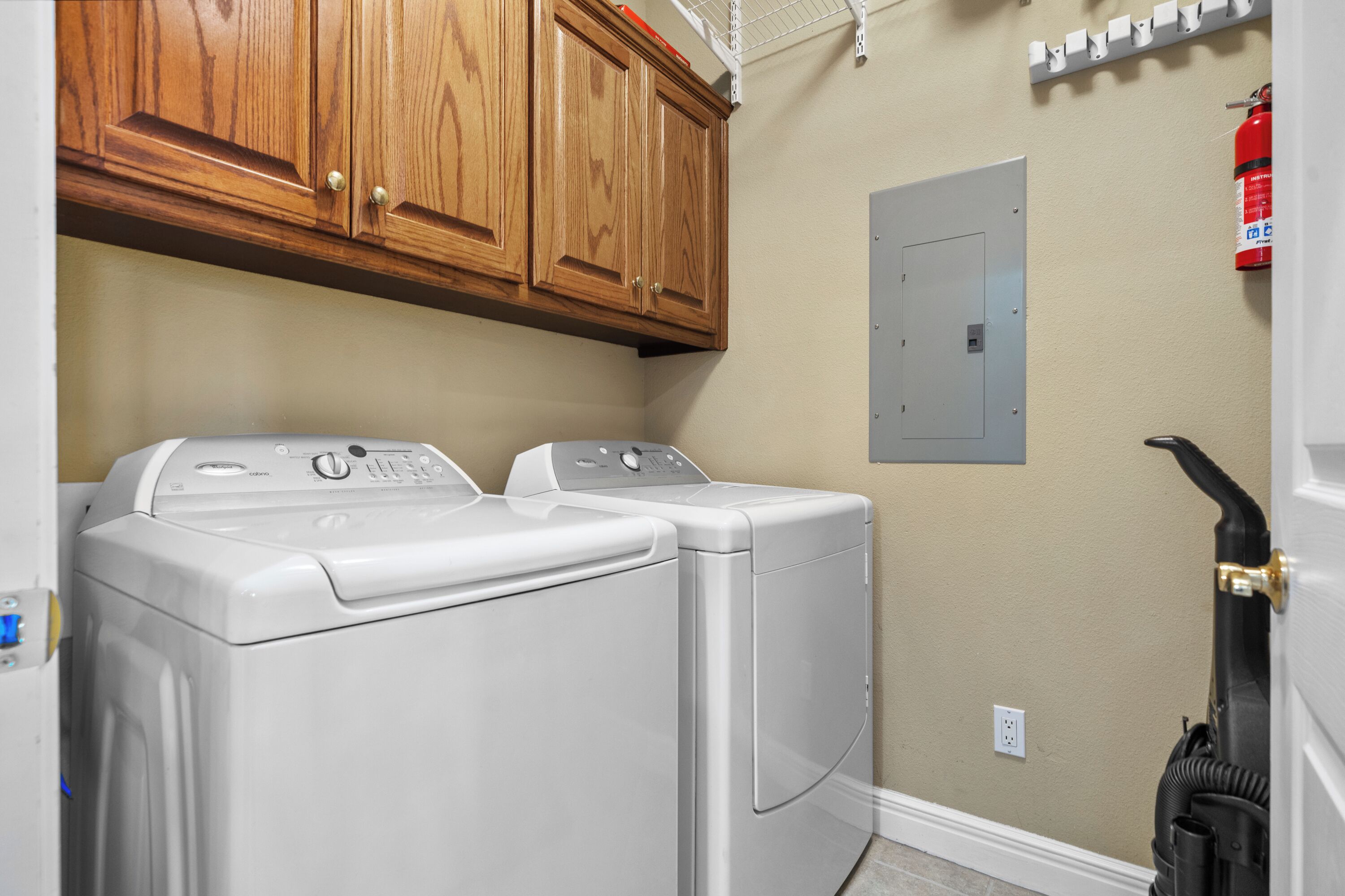 A laundry area with two front-loading washing machines, wooden cabinets above, a fire extinguisher on the wall, and a door with a hose attached.