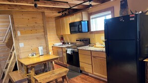 A kitchen with wooden cabinets, a black refrigerator, a microwave, and a stove. There is a wooden bench and a small table with a paper towel holder. - Authentic Fairbanks log house with bathroom, kitchen, and screened front porch (Fairbanks)