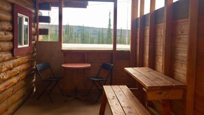A wooden cabin with a small dining area featuring a table and two chairs, a bench, and a window offering a view of a forested landscape. - Authentic Fairbanks log house with bathroom, kitchen, and screened front porch (Fairbanks)