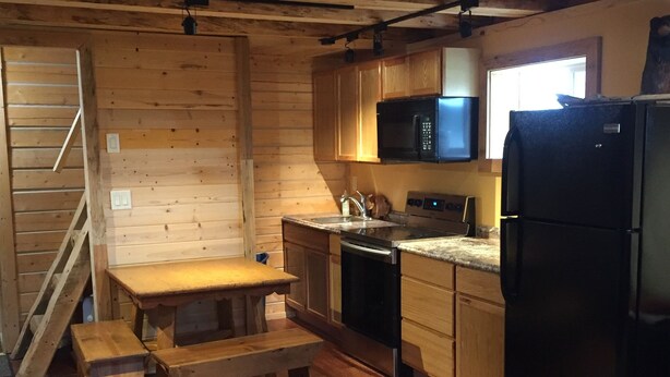 A compact kitchen with wooden cabinets, a microwave, a refrigerator, and a dining area with a bench. - Authentic Fairbanks log house with bathroom, kitchen, and screened front porch (Fairbanks)