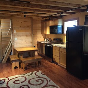 A compact kitchen with wooden cabinets, a microwave, a refrigerator, and a dining area with a bench. - Authentic Fairbanks log house with bathroom, kitchen, and screened front porch (Fairbanks)