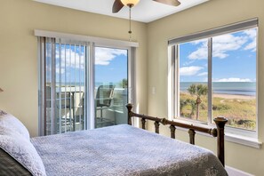 Un dormitorio con cama, ventilador de techo, un ventanal con vistas a la playa y puerta corrediza de vidrio.