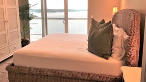 A bedroom with a large bed, a view of a body of water through a sliding door, a potted plant, and a lamp on the nightstand.