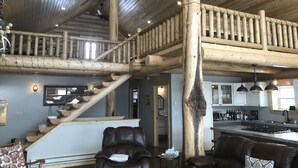 A spacious living area with a high ceiling, wooden beams, a staircase, and a kitchen area visible in the background.