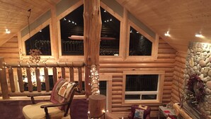 A log cabin interior with a cozy seating area, a stone fireplace, and a large window overlooking the night sky.
