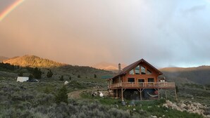 A wooden cabin with a balcony, surrounded by a mountainous landscape and a rainbow.