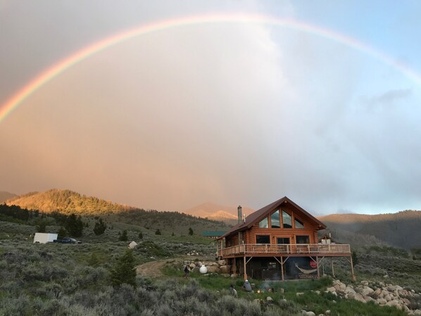 A wooden cabin with a balcony, surrounded by a mountainous landscape and a rainbow.