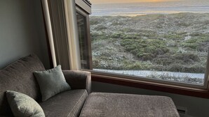 Ein Zimmer mit Blick auf eine Küstenlandschaft bei Sonnenuntergang, ein graues Sofa und ein Fenster mit Jalousien.