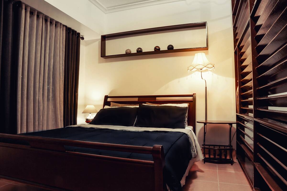 A hotel room with a wooden bed, a bedside table with a lamp, a shelf with decorative items, and a wardrobe with a mirror.