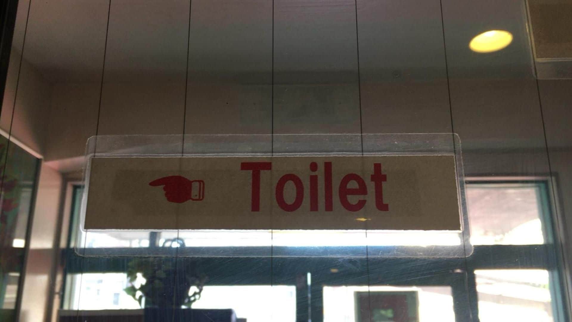 Señal con la palabra 'Toilet' e icono de una mano apuntando, indicando la ubicación de un baño.