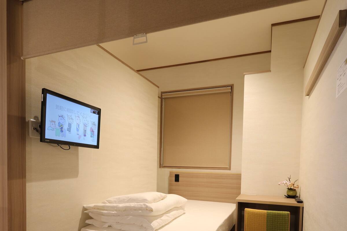 A hotel room with a bed, a TV mounted on the wall, a small table with a vase, and a carpeted floor.