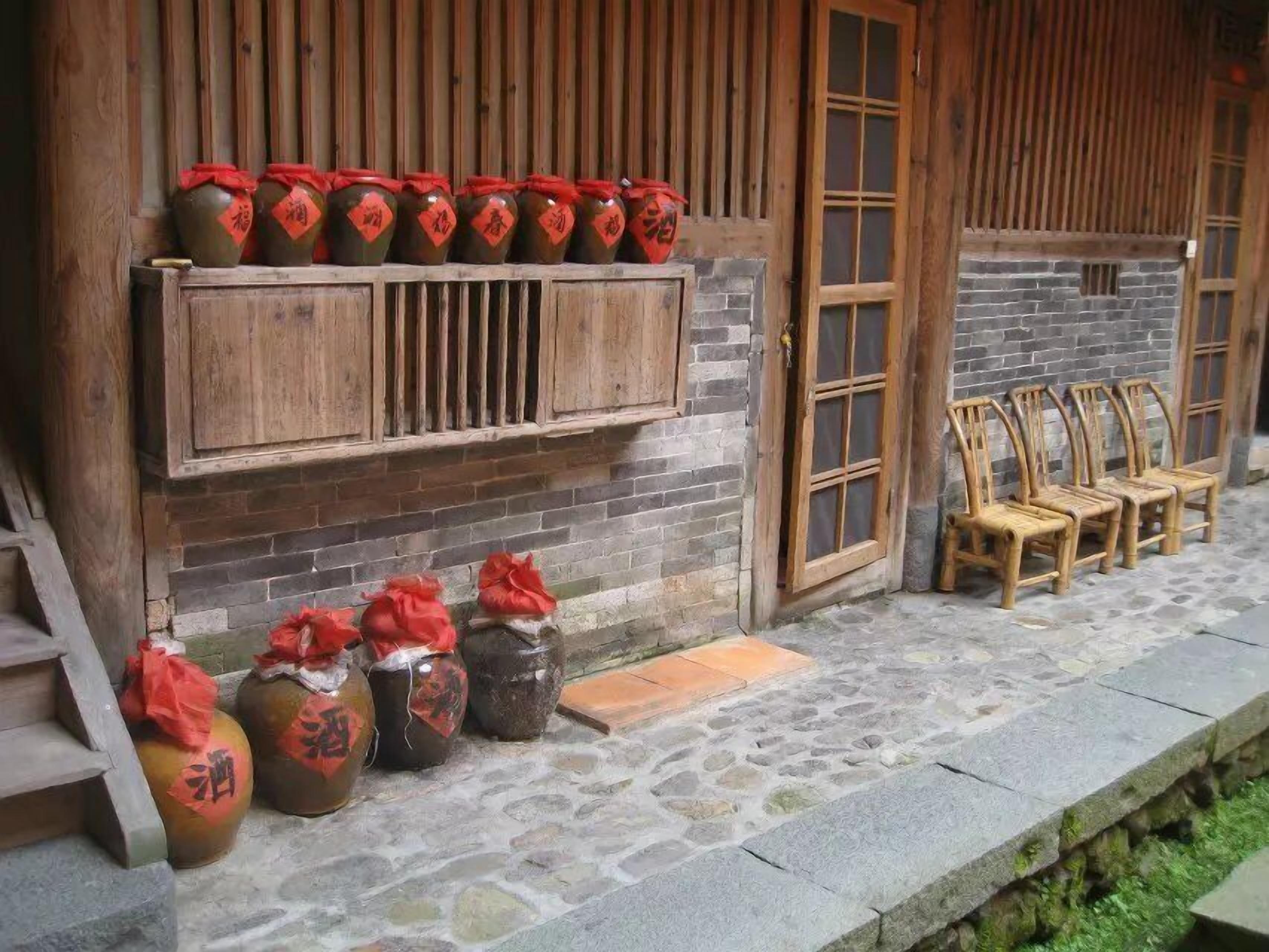 Un bâtiment traditionnel en bois, doté d’une entrée pavée de pierres, de bancs en bois et de pots portant des étiquettes rouges.