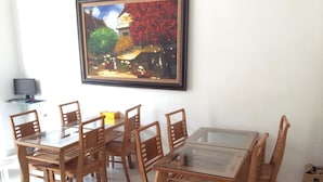 Un coin repas avec une table à plateau en verre et des chaises en bois, un tableau encadré accroché au mur et un téléviseur posé sur un meuble.