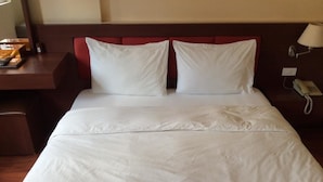 Un lit bien fait, avec du linge de lit blanc, une table de chevet en bois sur laquelle se trouve une lampe, et un téléphone.