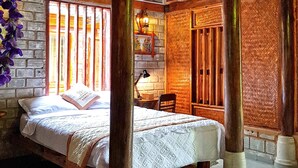 Quarto com cama, escrivaninha e cadeira. Há vigas de madeira e uma parede de tijolos.