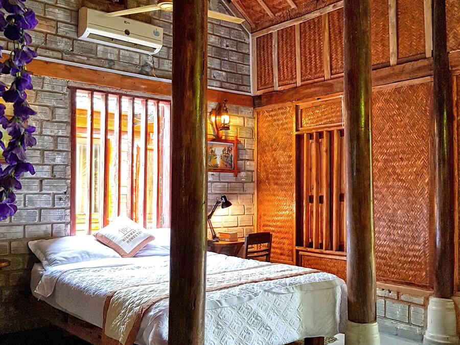 Una habitación con una cama, un escritorio y una silla. Se observan vigas de madera y una pared de ladrillo.