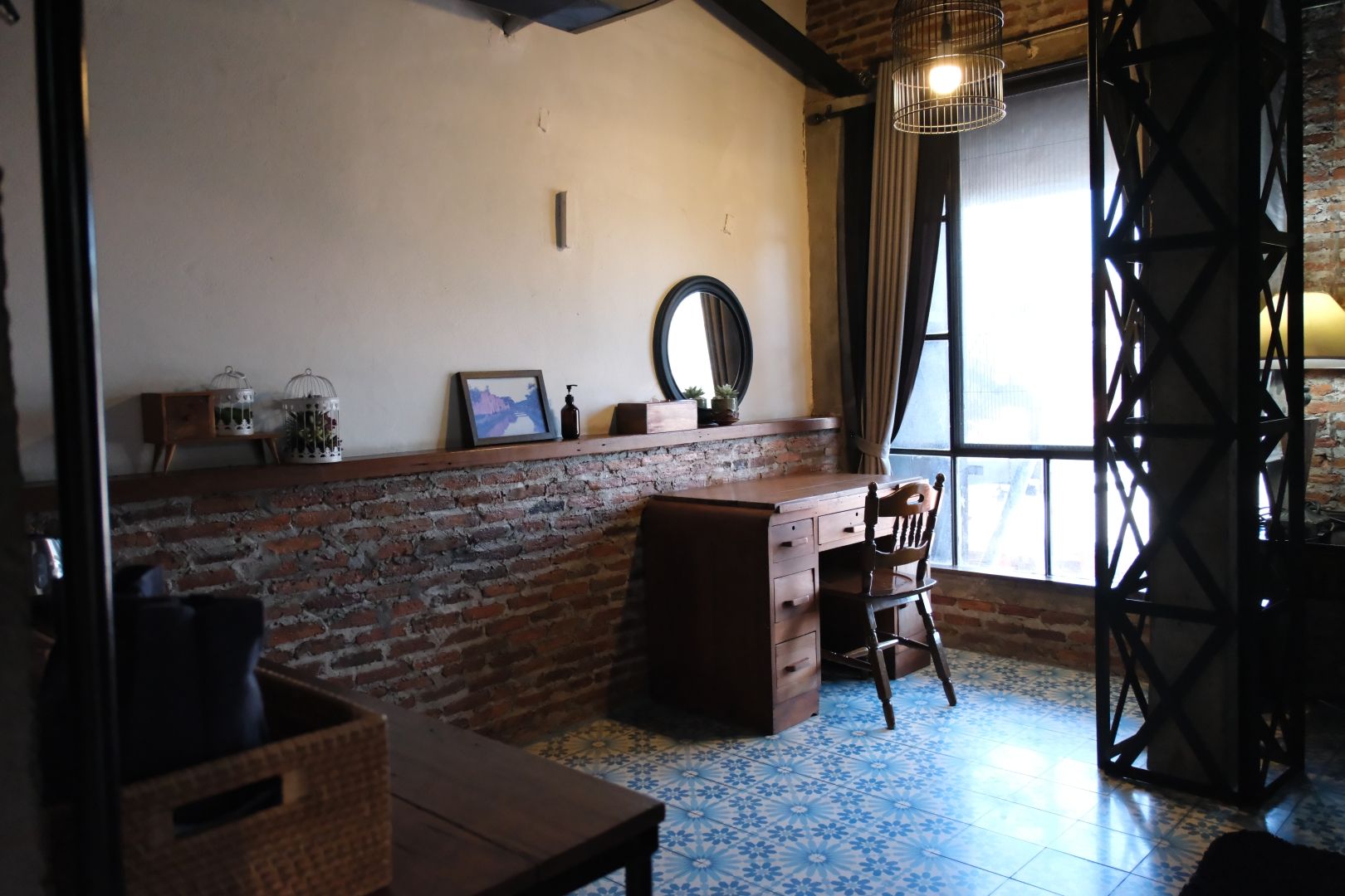 A room with a brick wall, a desk with drawers, a chair, a window with curtains, and a patterned floor.
