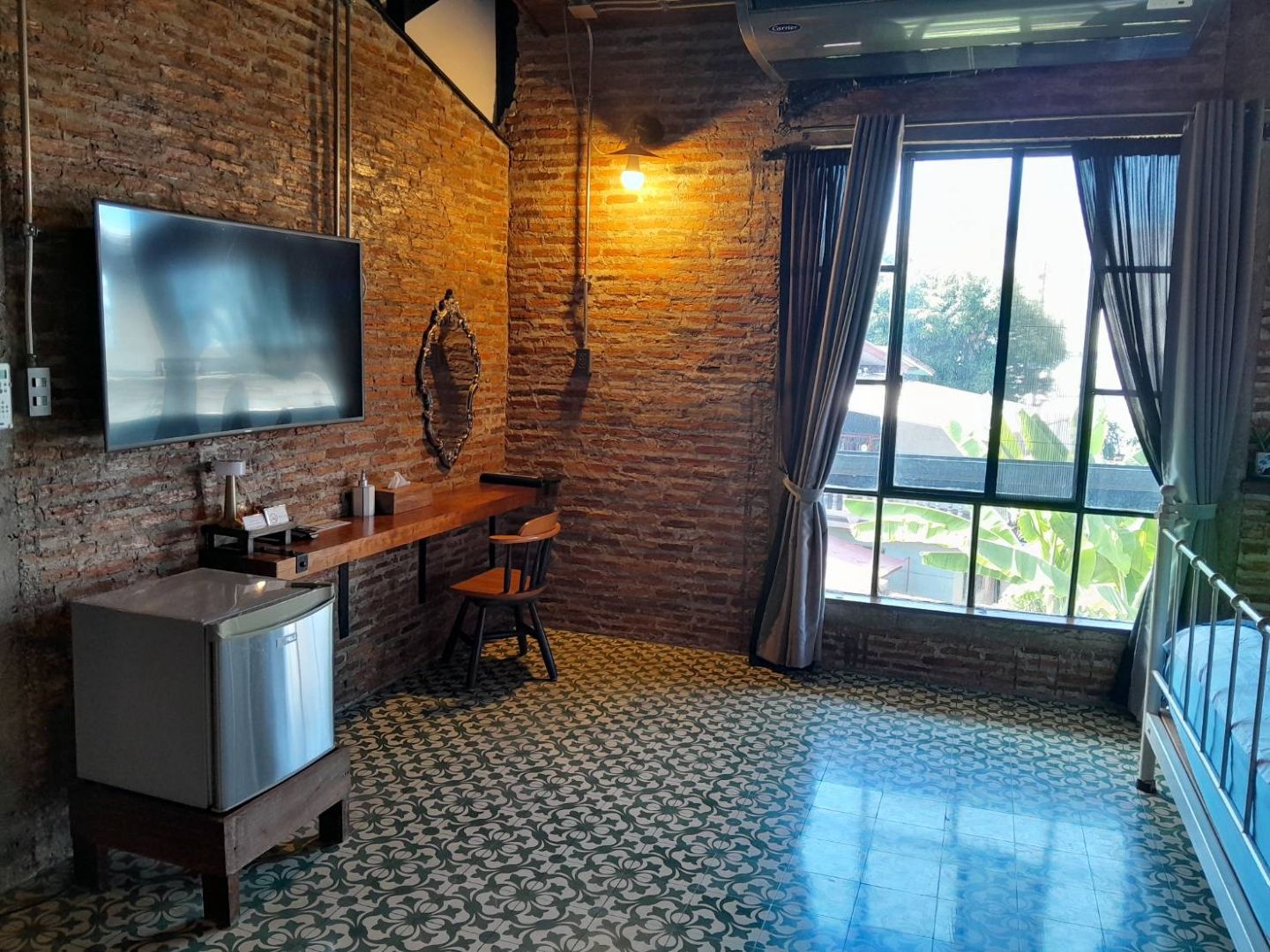 A room with a brick wall, a flat-screen TV mounted above a wooden table, a small refrigerator, and a patterned floor.